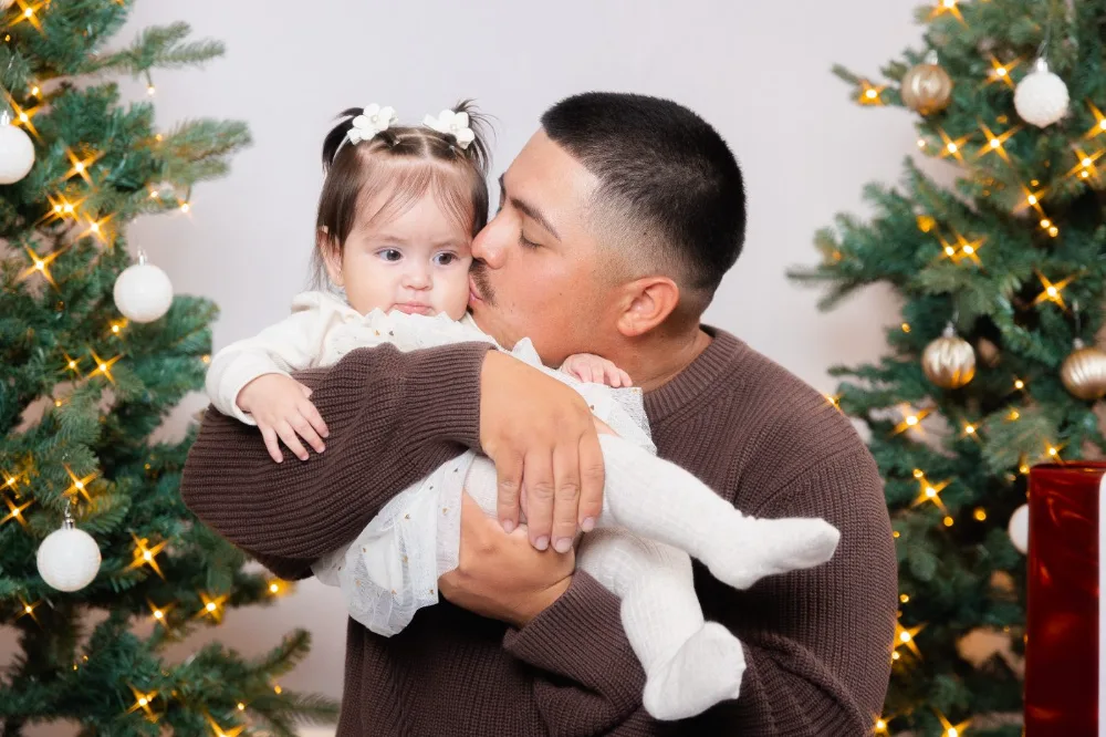 Dad kissing his baby in front of a lit Christmas tree